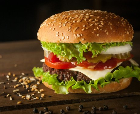 A sesame seed hamburger bun with a beef patty, lettuce, tomato, onion, pickles, cheese, and sauce sits on a dark wooden surface with scattered grains and peppercorns.
