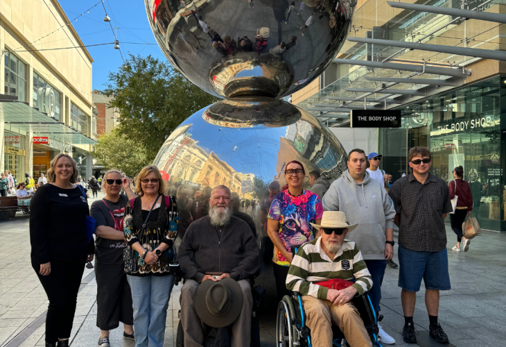 A group of people posing for a photograph in front of a large reflective sculpture in an urban outdoor setting.