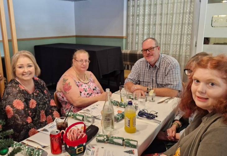 Five people sitting at a table with food and drinks, looking at the camera. Holiday-themed decorations are on the table.