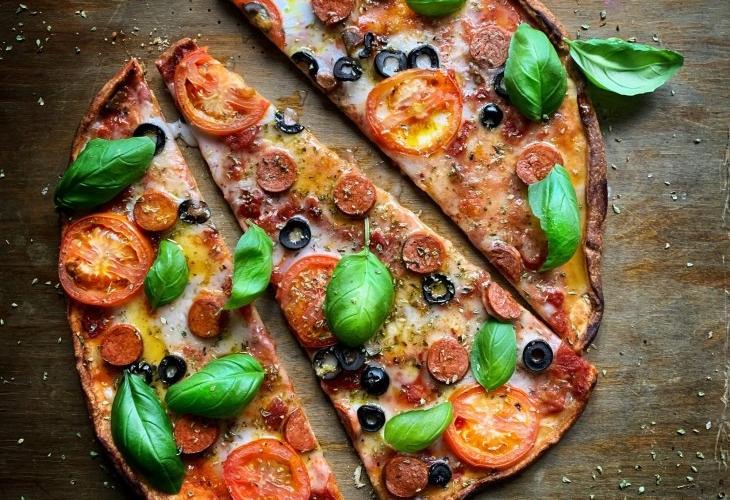 A thin-crust pizza topped with pepperoni, tomato slices, black olives, cheese, and fresh basil, cut into four slices on a dark wooden surface sprinkled with herbs.