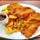 A plate with breaded schnitzel, golden roasted potatoes, a lemon wedge, and a small bowl of red sauce on a wooden table. A fork and knife rest on a napkin nearby.