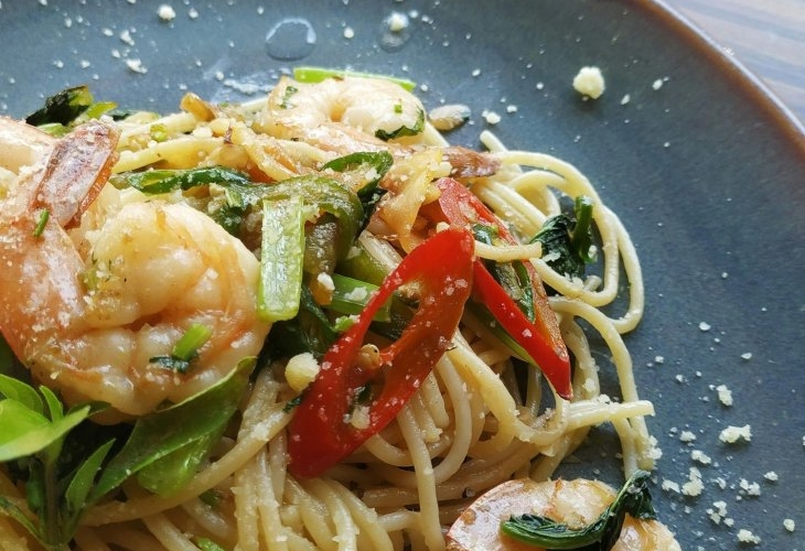 A plate of spaghetti with shrimp, sliced red chili, greens, and grated cheese on a dark dish.