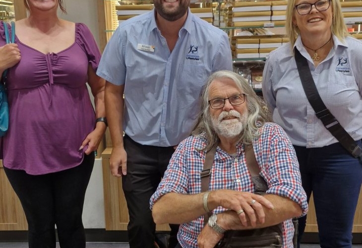 Four people posing indoors, with two standing on each side of a man sitting in a wheelchair. They are in a room with shelves in the background.