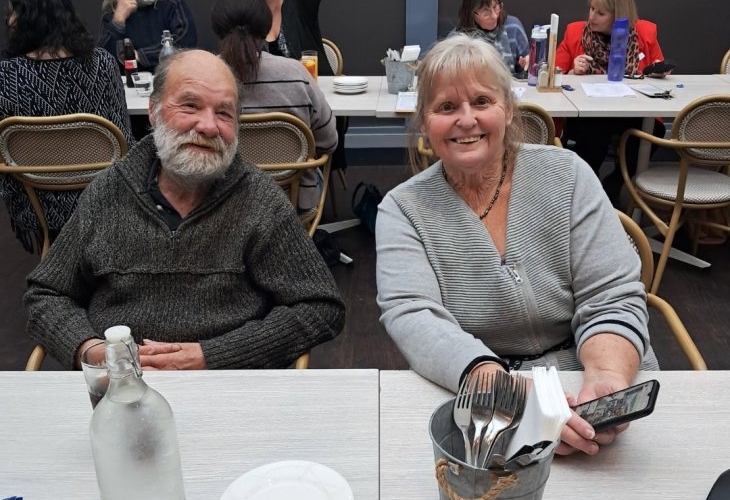 Two elderly people sit at a table in a restaurant, smiling at the camera. Other diners are seen in the background, engaged in conversation.