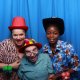 Three people pose in front of a blue curtain wearing colorful hats and balloon accessories, smiling at the camera.