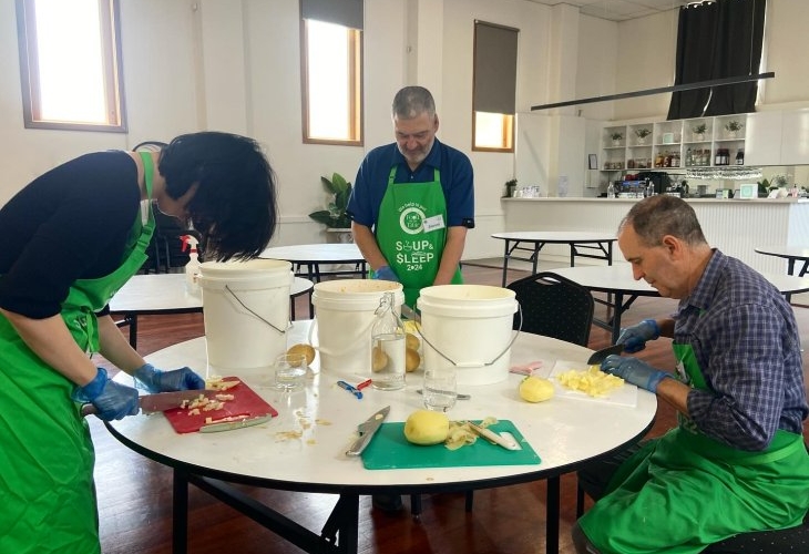 Three people in green aprons are peeling and chopping potatoes at a round table in a spacious room with buckets and kitchen supplies.