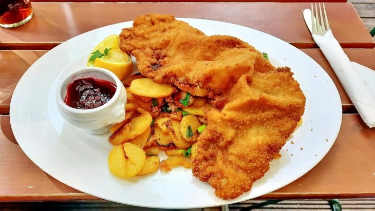 A plate with breaded schnitzel, golden roasted potatoes, a lemon wedge, and a small bowl of red sauce on a wooden table. A fork and knife rest on a napkin nearby.