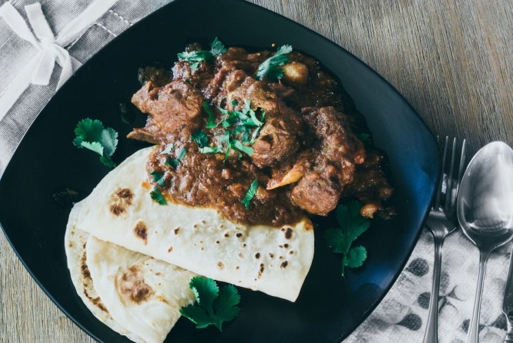 A plate with curry topped with cilantro, served with two flatbreads, on a wooden table with a fork and spoon on the side.