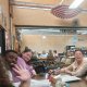 A group of people sitting at a table in a casual indoor setting. One person waves at the camera. The background includes other tables and an umbrella decor.