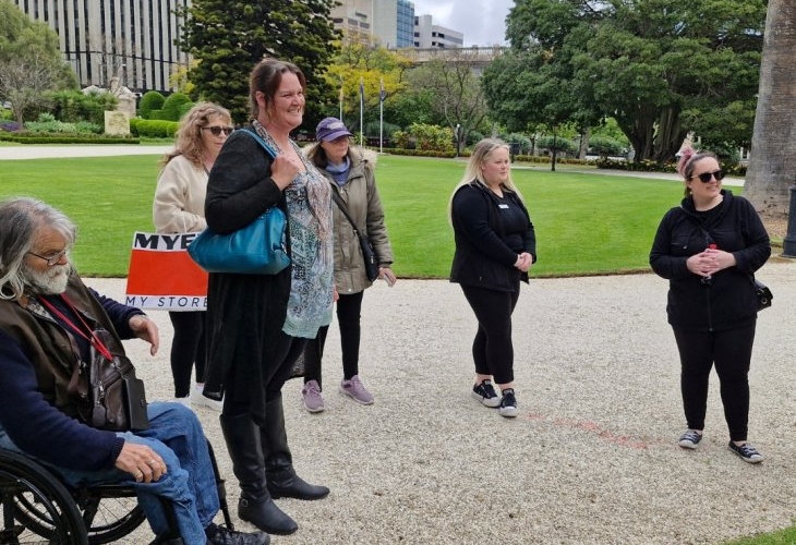 A group of people, including one in a wheelchair, stand on a gravel path in a park with trees and buildings in the background.