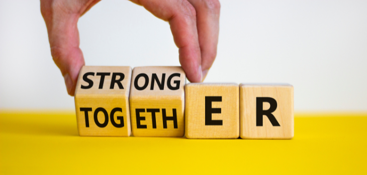 White and yellow background with 4 wooden cubes. One side says Strong and the other Togeth. The letters E and R are on the last two blocks spelling Stronger Together