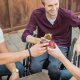 Three men, one in a wheelchair, smile as they clink glasses together in a toast while seated outdoors.