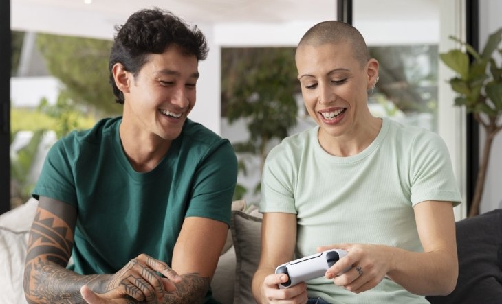 Two people sitting on a couch, smiling and interacting with a video game controller in a bright, modern living room.
