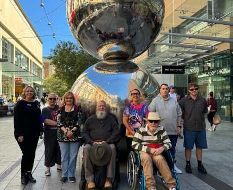 A group of people posing for a photograph in front of a large reflective sculpture in an urban outdoor setting.