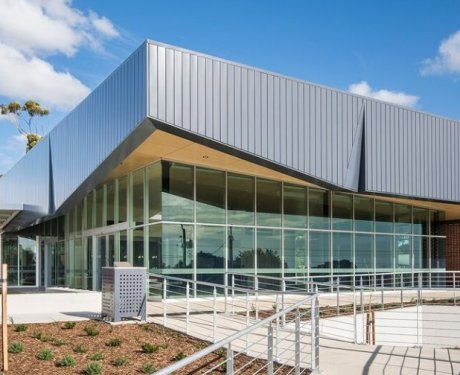 Modern building with large glass windows and metal cladding, surrounded by a landscaped area and a ramp leading to the entrance, under a blue sky with scattered clouds.