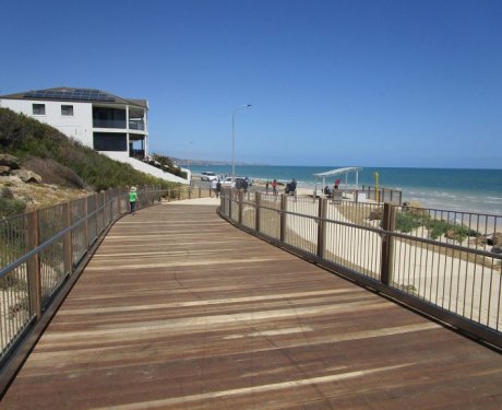 Wooden boardwalk leading to a beach with people in the distance, a building on the left, and clear blue sky.