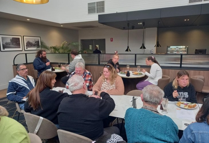 A group of people sitting around tables in a restaurant, eating and talking. Some are looking at menus, while others are engaged in conversation or enjoying their meals.