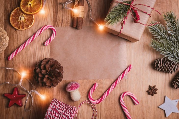 A festive arrangement on a wooden surface with candy canes, a wrapped gift, dried orange slices, pinecones, and holiday lights.