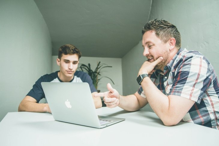 Two men sit at a table looking at a laptop. One man gestures towards the screen while the other watches attentively.