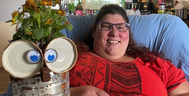 A female NDIS participant sitting in an arm chair with a vase of flowers on a table with jam jar lids, bottle tops and googly eyes glued to the vase to make it look like an owl