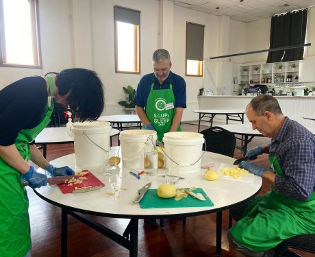 Three people in green aprons are peeling and chopping potatoes at a round table in a spacious room with buckets and kitchen supplies.