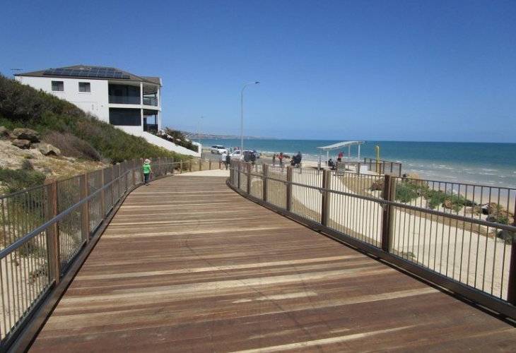 Wooden boardwalk leading to a beach with people in the distance, a building on the left, and clear blue sky.