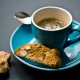 A blue cup of espresso on a matching saucer with a spoon, accompanied by a biscotti and sugar cubes on a dark table.