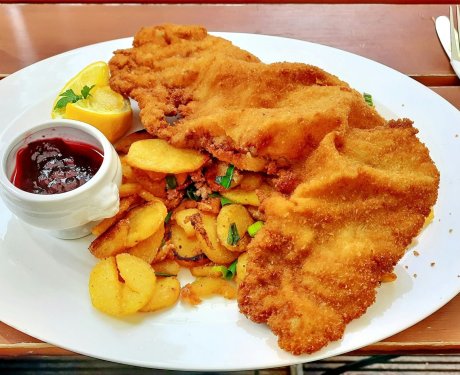 A plate with breaded schnitzel, golden roasted potatoes, a lemon wedge, and a small bowl of red sauce on a wooden table. A fork and knife rest on a napkin nearby.