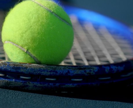 A close-up of a tennis ball resting on a blue tennis racket, with the focus on the ball and stringed surface of the racket.