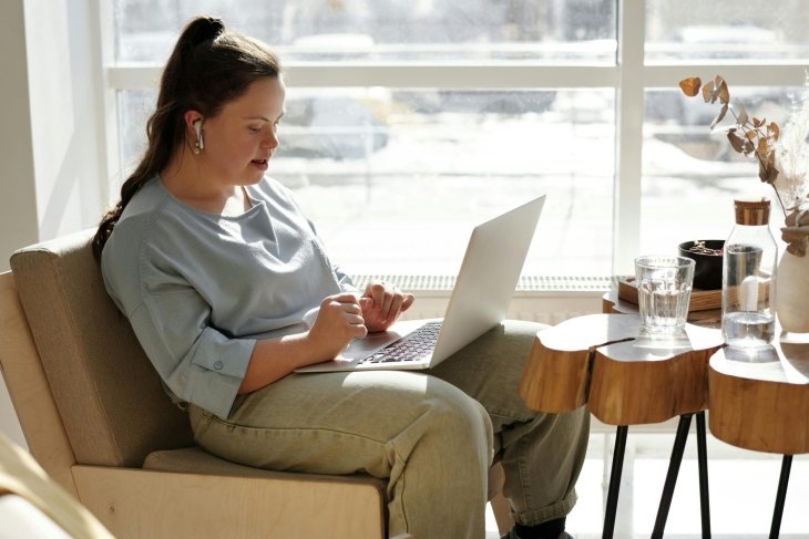 Woman sitting on a chair, typing on a laptop, with earphones in. A table with glasses and decorative items is beside her, and sunlight streams through a large window.