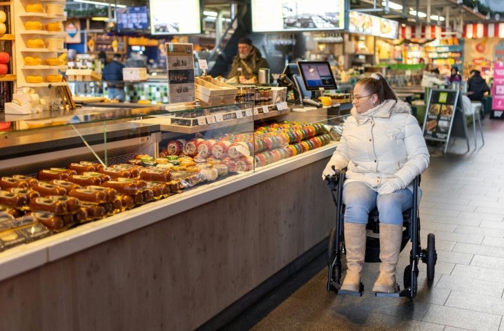 A woman in a wheelchair is shopping in a market, looking at an array of food items displayed in a glass case. The market is indoors, with various stalls and displays in the background.