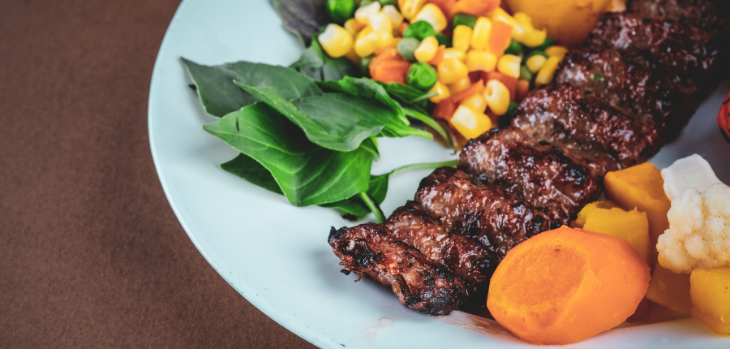 A white plate with cooked beef, peas, corn, carrots and leafy greens