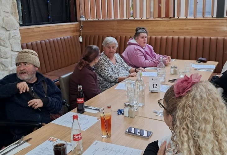 A group of people sit around a rectangular table in a restaurant, engaged in conversation. Various drinks and menus are visible on the table.