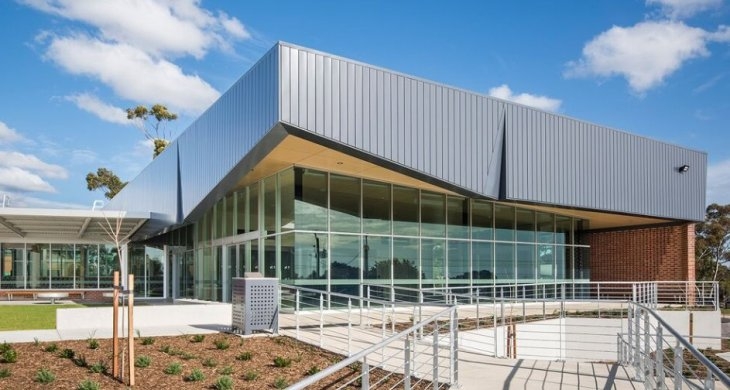 Modern building with large glass windows and metal cladding, surrounded by a landscaped area and a ramp leading to the entrance, under a blue sky with scattered clouds.