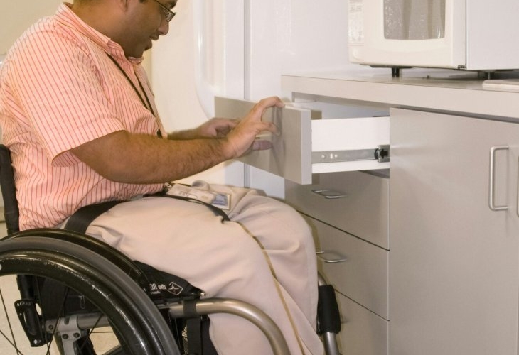 A man in a wheelchair accessing a microwave placed on a lower cabinet in an accessible kitchen.