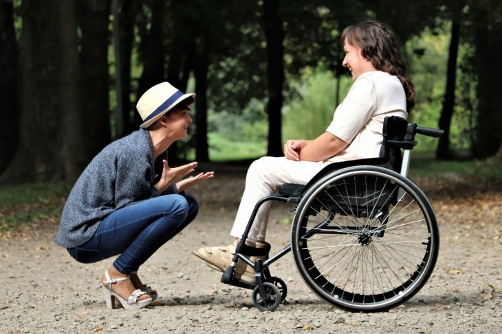 A woman in a hat squats and converses with a smiling woman in a wheelchair on a park pathway surrounded by trees.