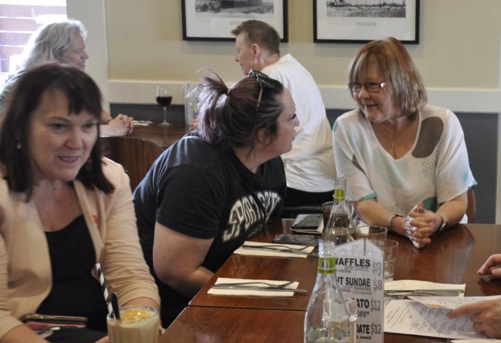 A group of people are seated around a table in a restaurant, engaged in conversation. Menus, drinks, and a bottle of water are on the table. Framed black-and-white photographs are on the wall in the background.