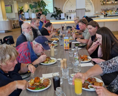 A group of people sitting at a long table eating a meal in a restaurant. Plates of food and drinks are on the table.