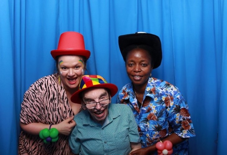 Three people pose in front of a blue curtain wearing colorful hats and balloon accessories, smiling at the camera.