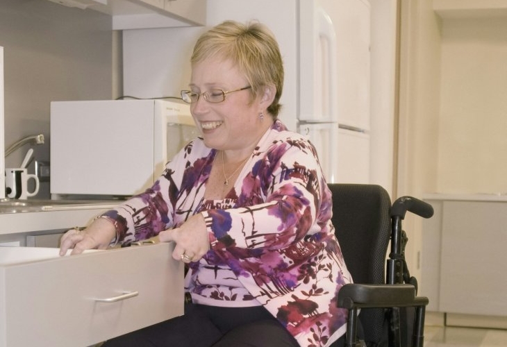 A woman in a wheelchair opens a drawer in a kitchen, smiling while engaged in the task.