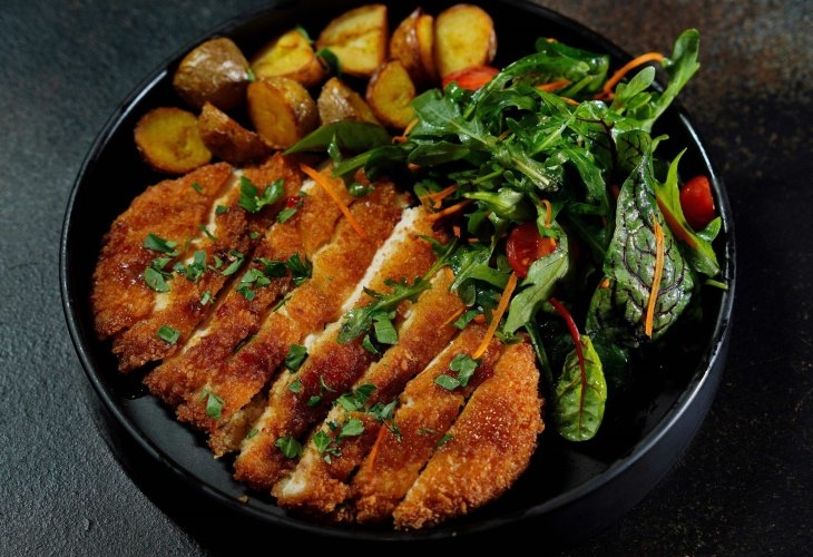 A black plate with sliced breaded chicken cutlet, roasted potatoes, and mixed green salad including arugula and cherry tomatoes.