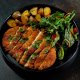 A plate with sliced breaded chicken, roasted potatoes, and a leafy green salad with cherry tomatoes.