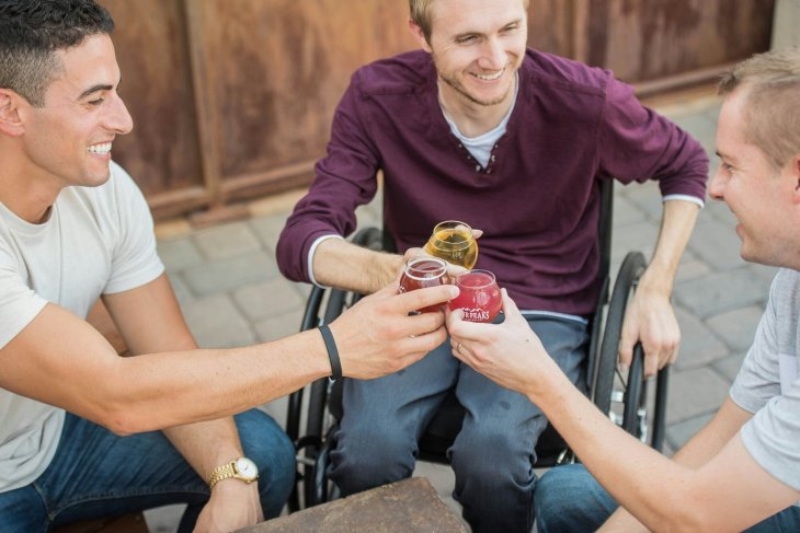 Three men, one in a wheelchair, smile as they clink glasses together in a toast while seated outdoors.