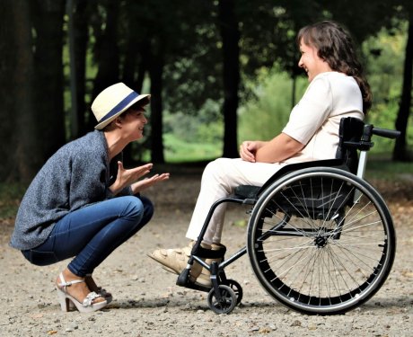 A woman in a hat squats and converses with a smiling woman in a wheelchair on a park pathway surrounded by trees.