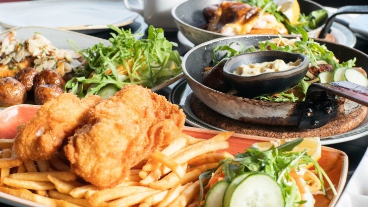 A table filled with various dishes, including fried fish and chips, salads with greens and cucumbers, and roasted potatoes, presented in pans and plates.