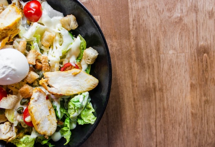 A bowl of Caesar salad with grilled chicken, cherry tomatoes, croutons, lettuce, and a poached egg on a wooden table background.