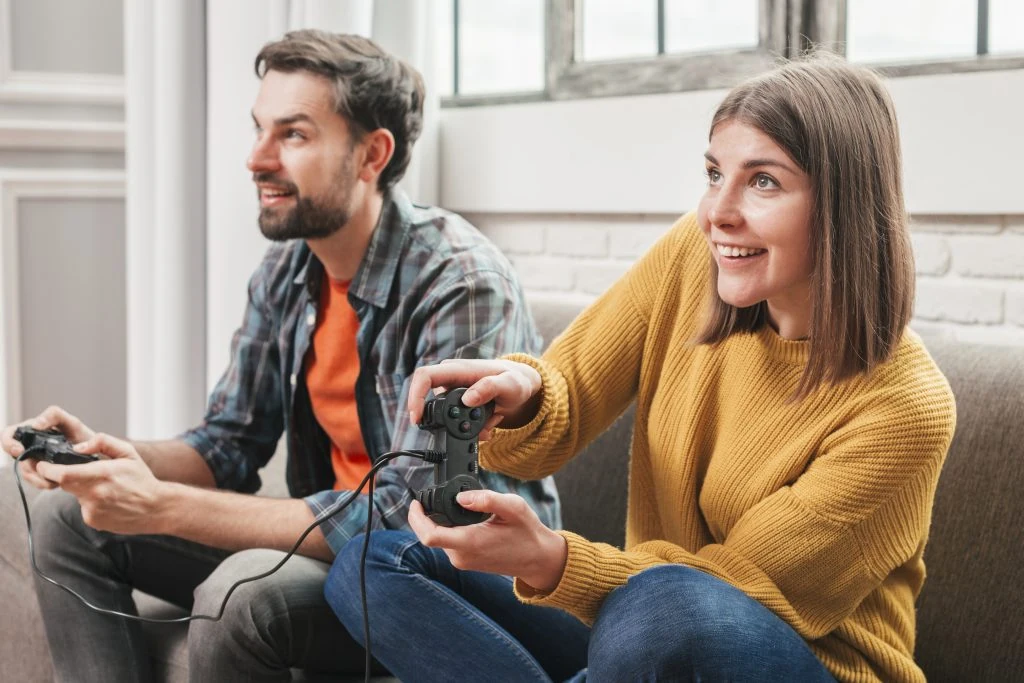 A man and a woman sit on a couch playing video games with wired controllers, both looking at a screen and smiling.
